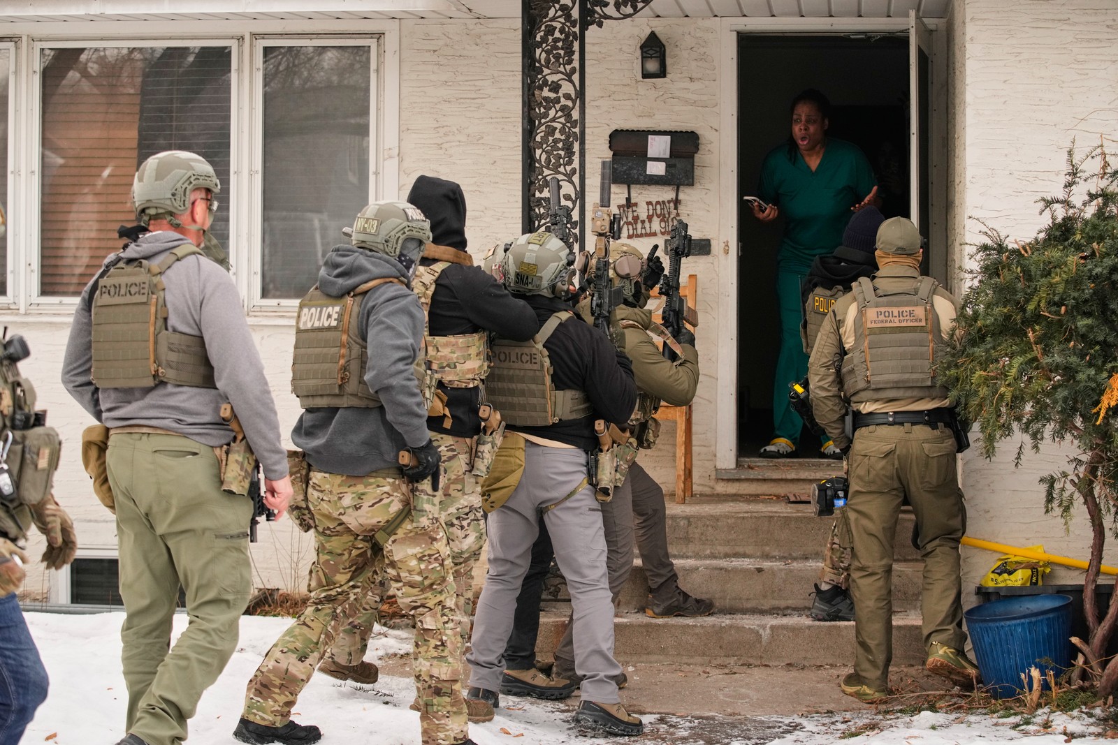 At least eight armed federal officers stand outside the open door of a house as a person inside gestures while speaking to them.
