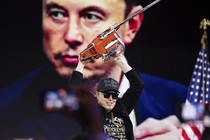 Photo of Elon Musk holding a chainsaw over his head onstage with a photo of himself projected on a screen behind him