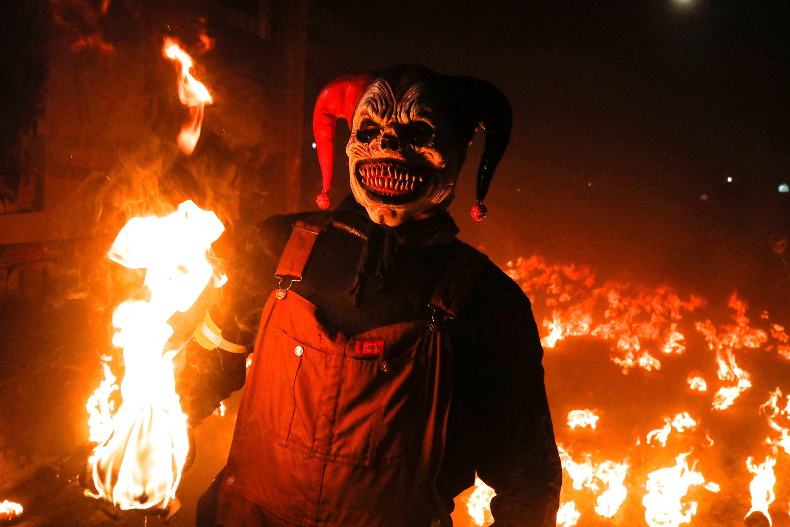 A person wearing a frightening clown mask poses in front of a fiery background.