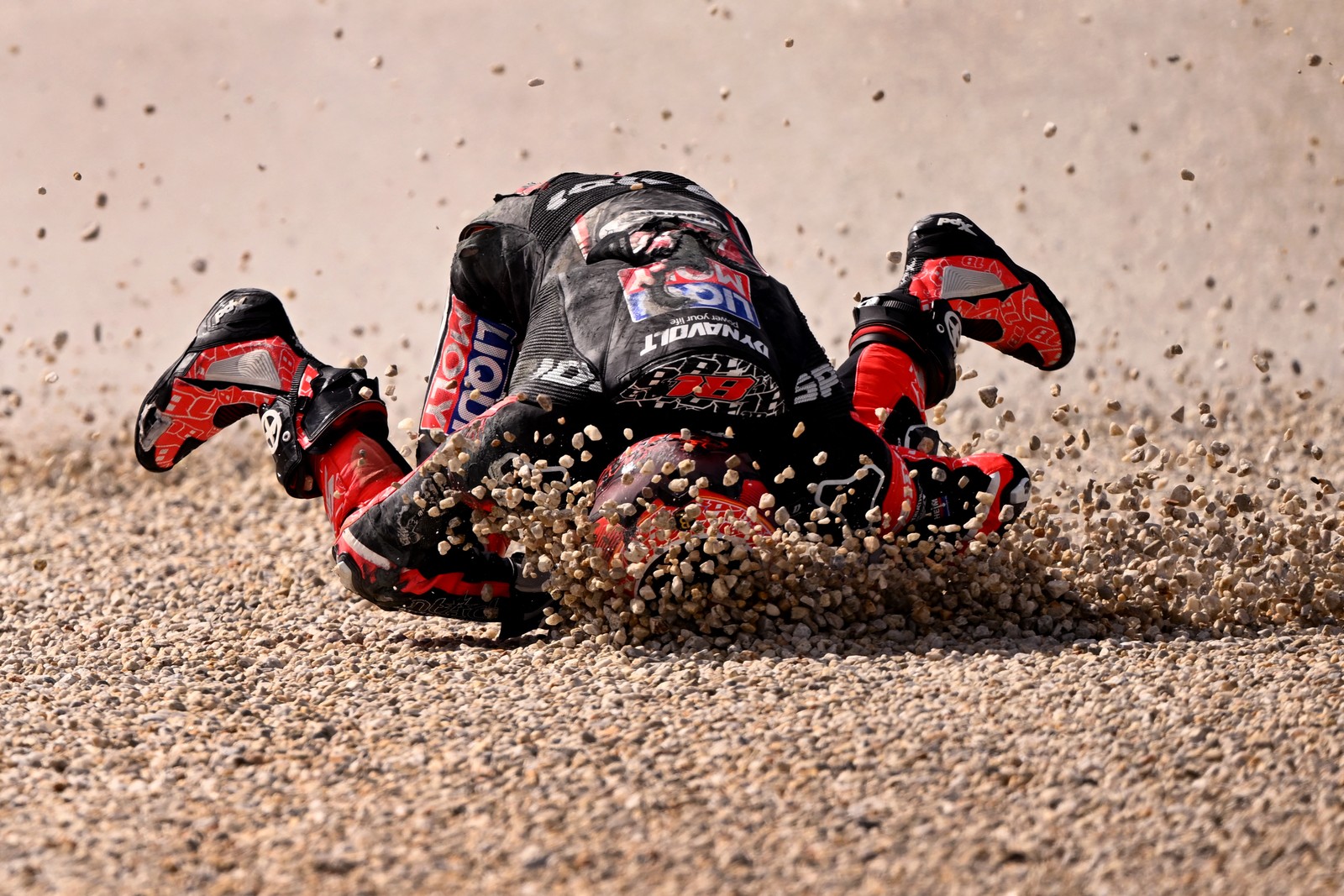 A motorcycle racer falls headfirst into a patch of gravel, their helmet sending bits of gravel flying.