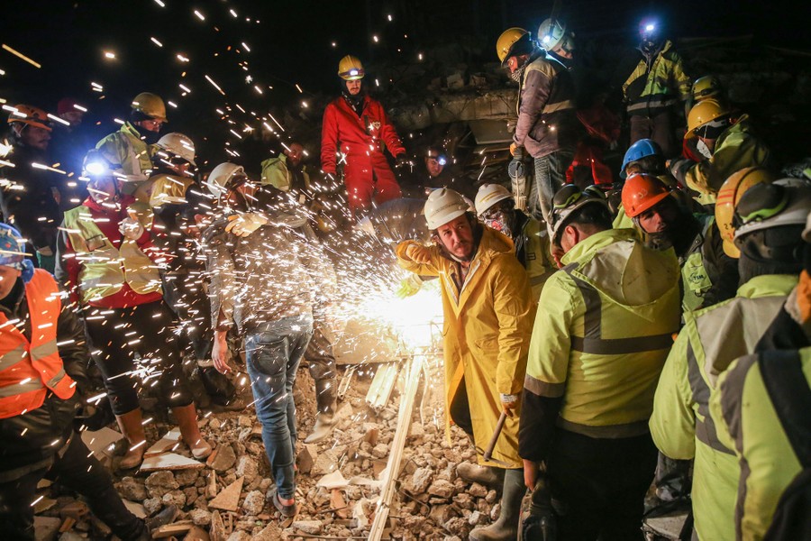 About a dozen rescue workers stand beside the rubble of a destroyed building, with sparks flying in the middle of the photo.