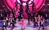 Ryan Gosling, dressed as Ken in a pink suit and gloves, sings into a microphone on the Oscars stage as several dozen men in black tuxedos dance behind him.