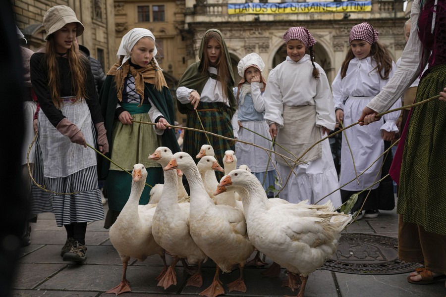 Girls in traditional clothing herd a small gaggle of geese in a city plaza.