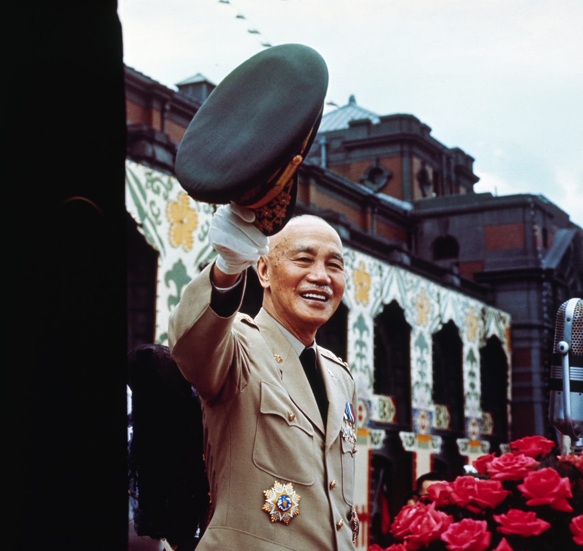 photo of man in military uniform smiling and waving hat with gloved hand, with bouquet of roses in foreground