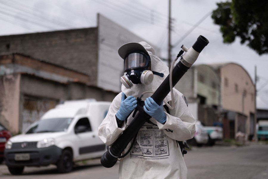 A person wearing protective gear and a mask carries a long tube used to fumigate an area.
