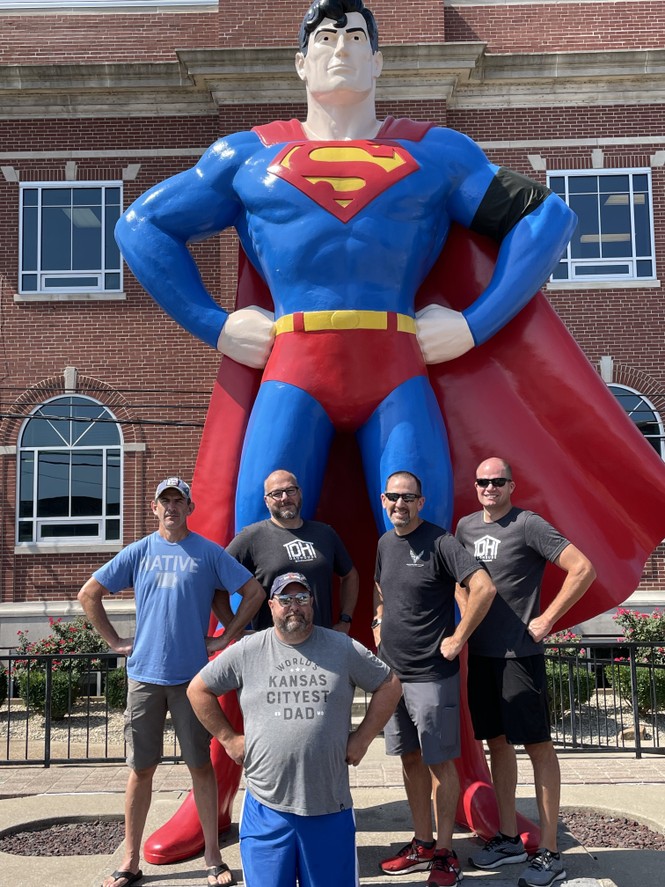 Five men stand with their hands on their hips, mimicking the pose of a giant Superman statue behind them