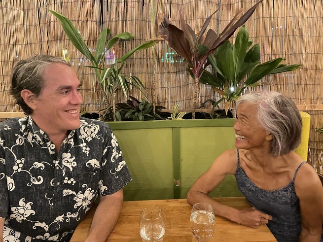 A middle aged man in a Hawaiian shirt sits across the table from an older woman wearing a sundress. They're smiling and leaning toward each other. There are potted plants in the background.