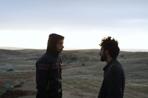 Two men, one in a hoodie and one with shaggy hair, stand in profile in a desert landscape.