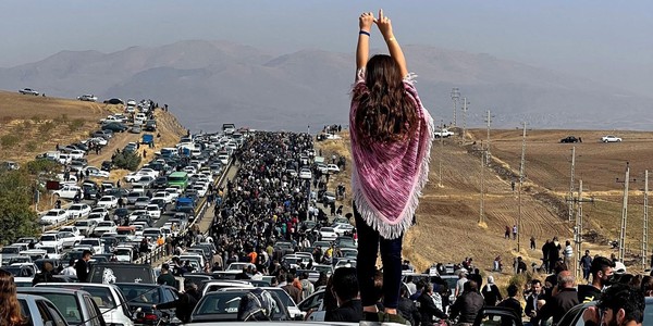 A woman holds her arms up, gesturing, while standing on a car among a large crowd of people marching past cars on a major road.