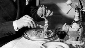 A black-and-white photograph of a torso in a suit and two hands swirling spaghetti against a spoon