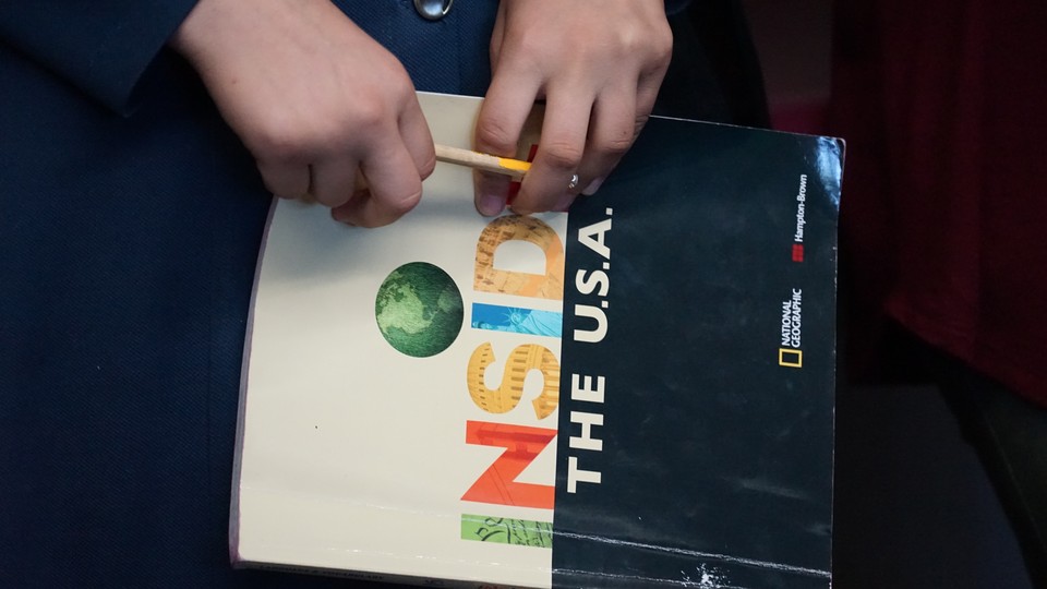 A student's hands hold a pencil in front of a book whose cover reads "Inside the USA"