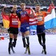 Three para-athletes wearing leg prostheses stand side by side, celebrating, draped in their countries' flags, inside a stadium.
