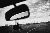 a black and white photo of the road and the cloudy sky seen out the front window of a car, the mirror taking up the front of the frame