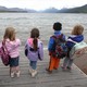 Five children holding backpacks look out at the expansive lake and mountains of Glacier National Park.