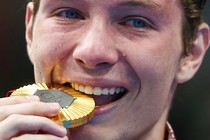 A close view of an athlete's face during a medal ceremony. He is playfully biting a gold medal, with tears falling from both eyes.