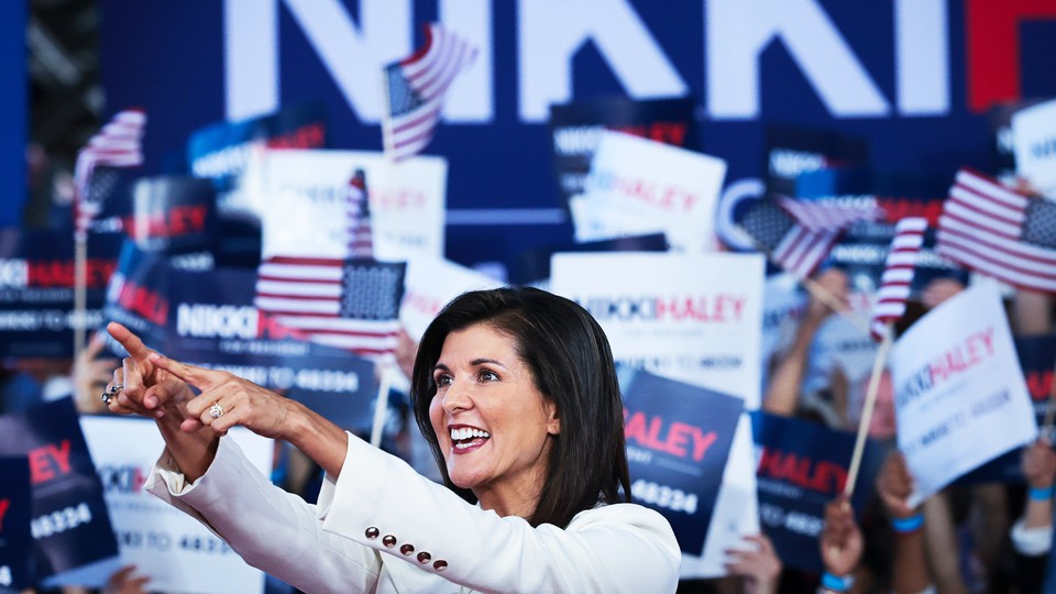 A photo of Nikki Haley in a white suit, pointing with both hands stretched forward, with a crowd behind her holding American flags and "Nikki Haley" signs