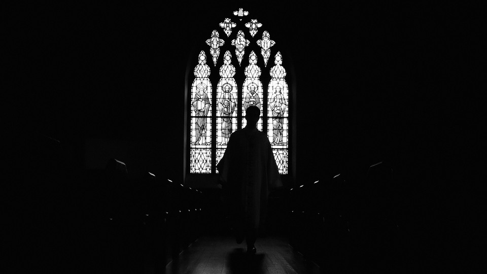 A black-and-white photo of the interior of an Episcopalian church. In the center of the image, a priest is backlit by sunlight coming through a stained glass window.