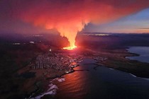 An aerial view of a coastal Icelandic town with an erupting volcanic fissure a short distance inland, just uphill from its houses