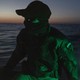 Photograph of a masked soldier on a small boat at dusk, with a green light shining on him