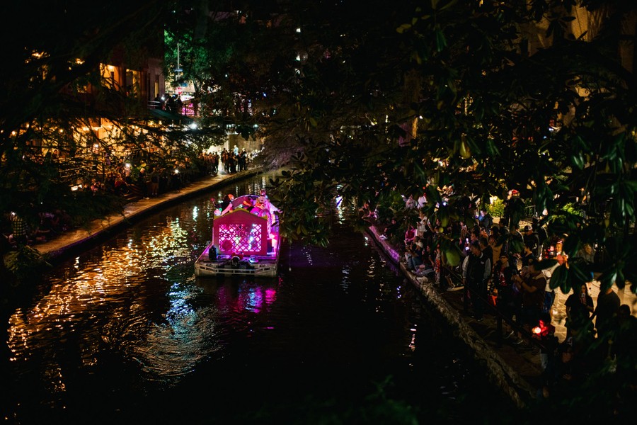 Many people gather on the banks of a canal, watching a colorfully-lit boat pass by, part of a night river parade.