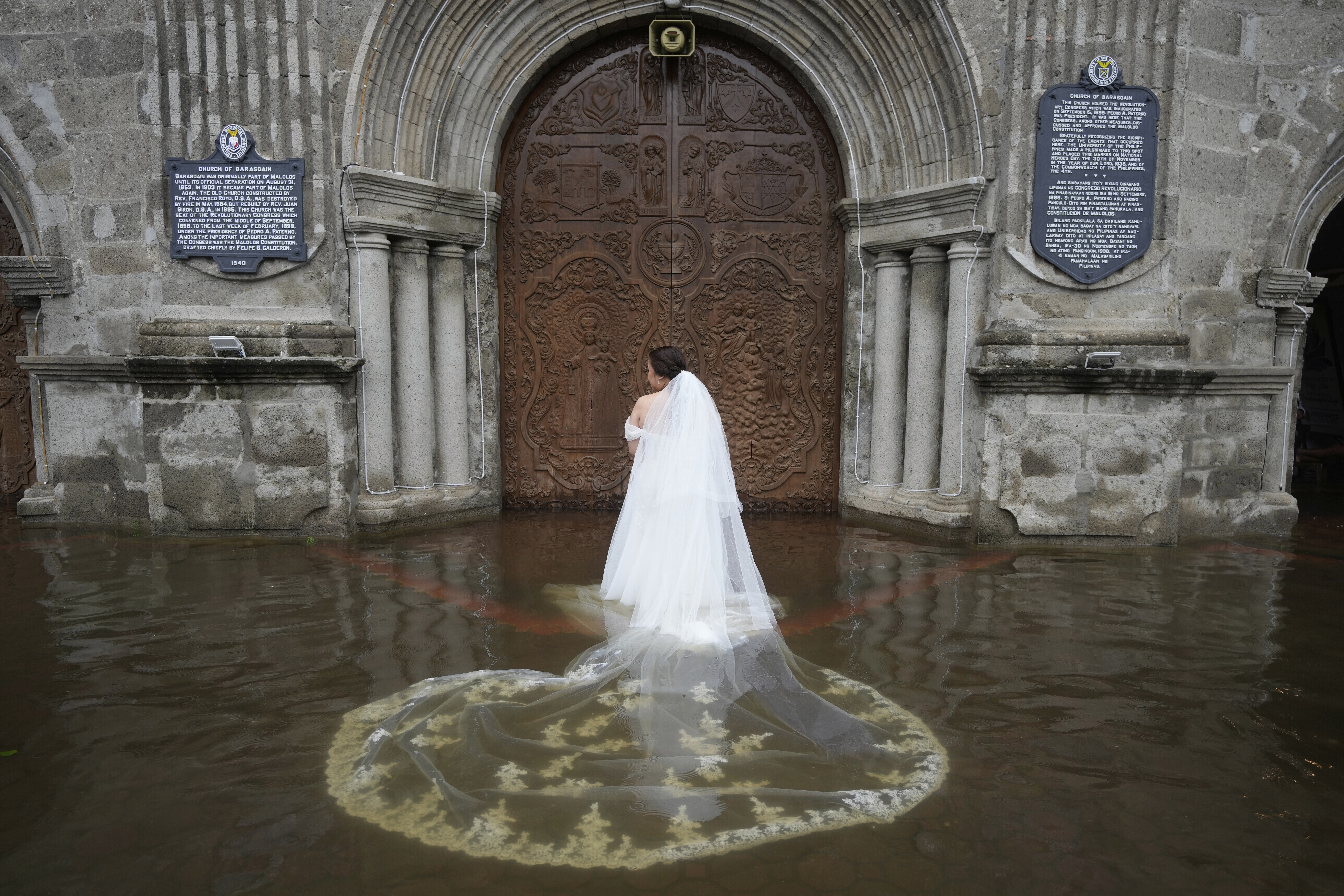 A bride with a long, flowing train, stands in foot-deep floodwater outside a church.