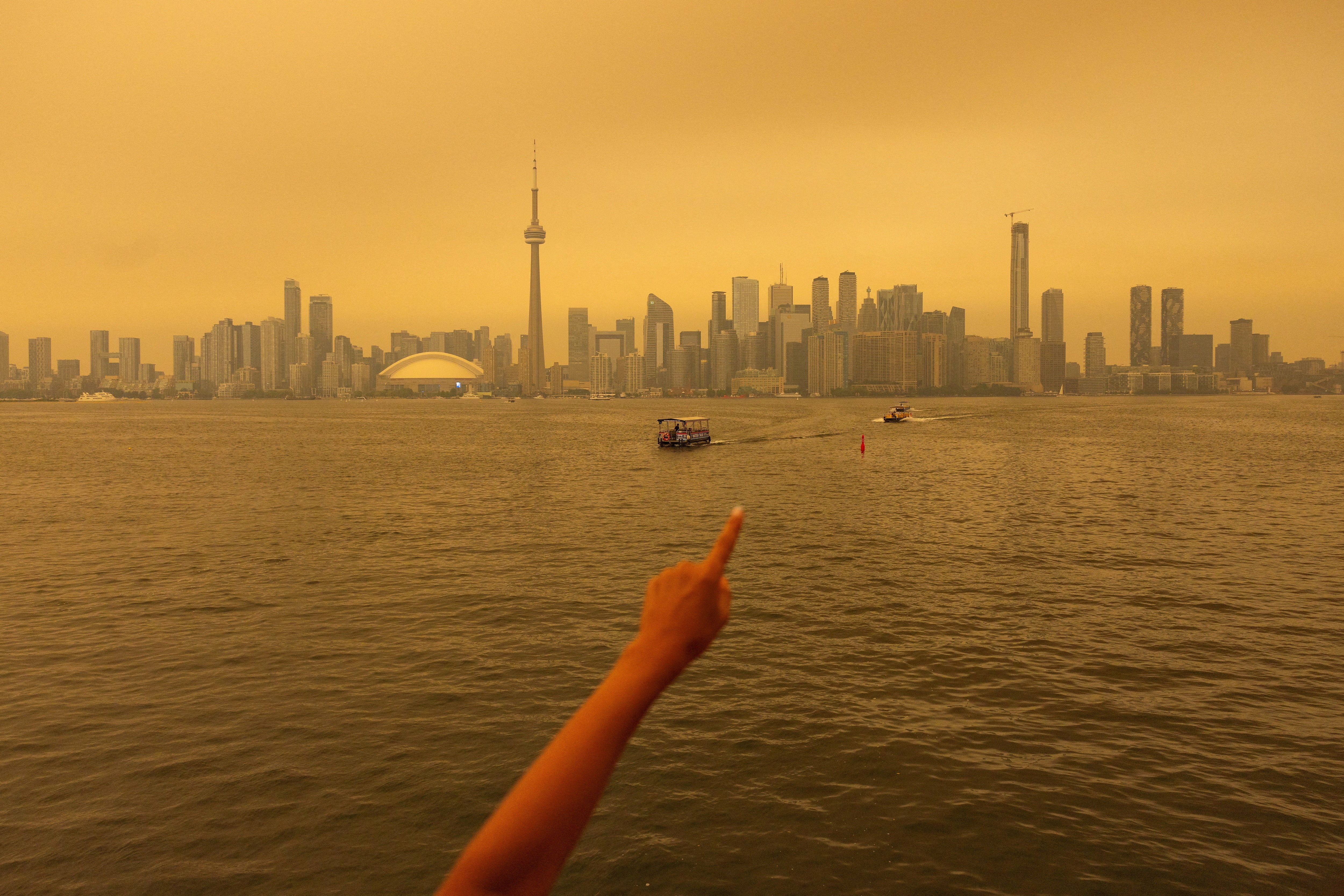 A person points to the Toronto skyline while they ride a ferry, under a yellowish smoky sky.