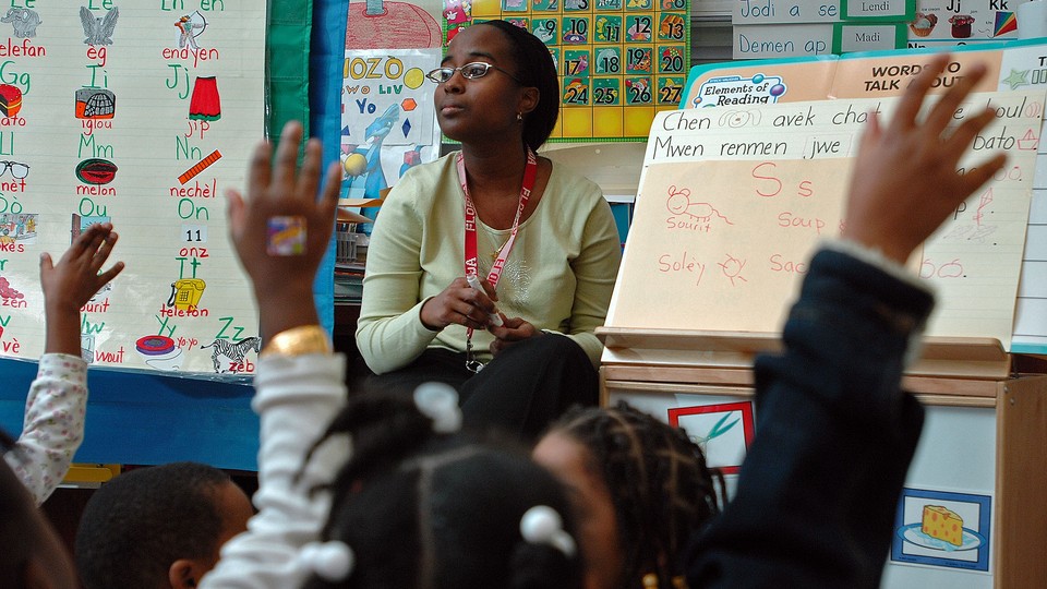 A teacher sits in front of her class. Students raise their hands.