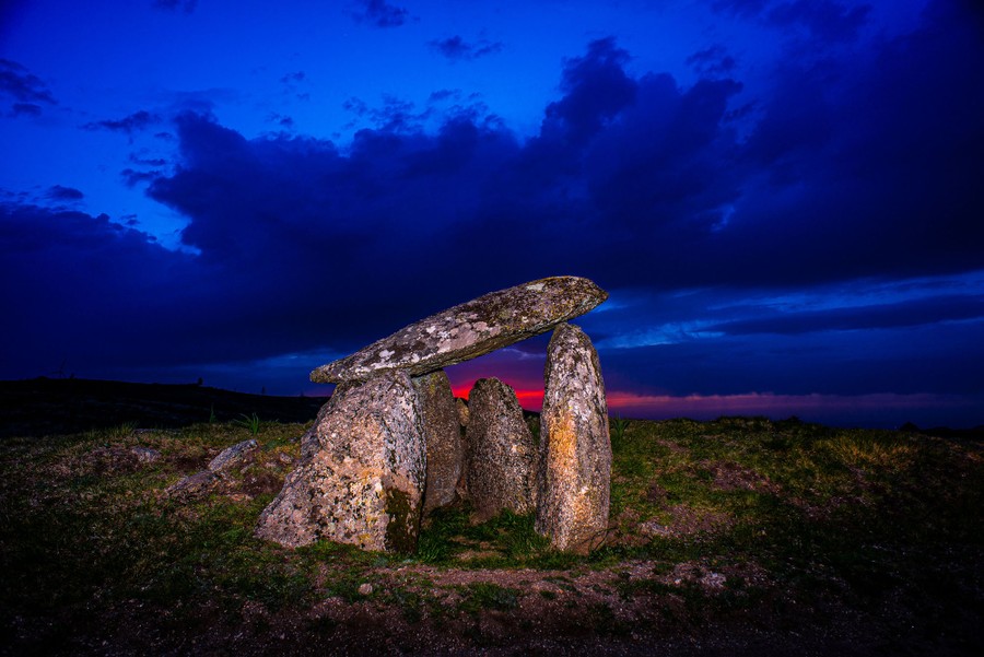 A large flat stone rests on top of several tall stones set in place to form a small chamber, on a grassy hill with a setting sun in the background.