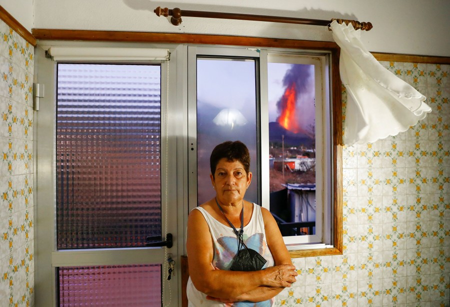 A woman stands in her home, with an erupting volcano visible through a window over her shoulder.