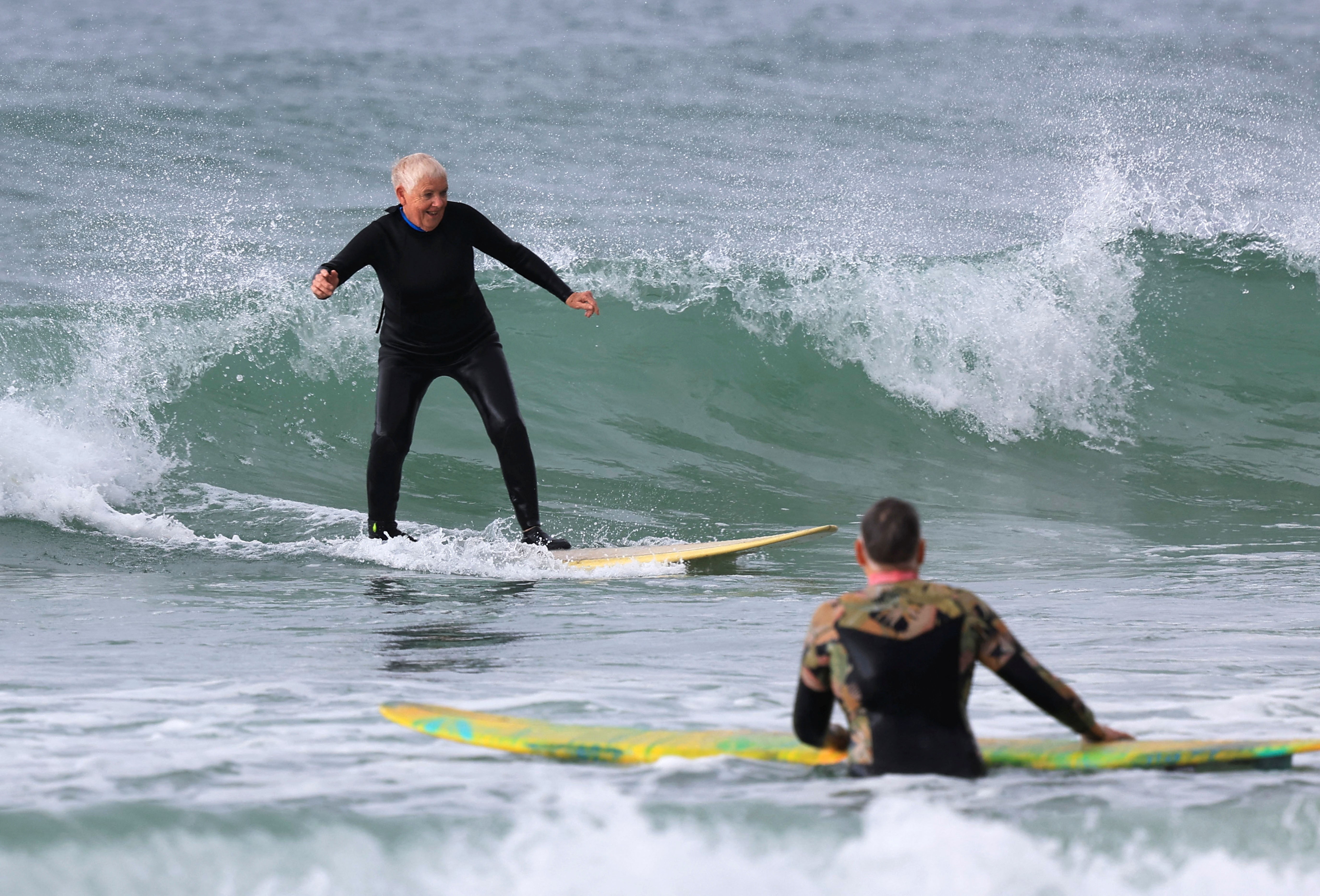 An older woman wearing a wetsuit rides a wave on a surfboard.