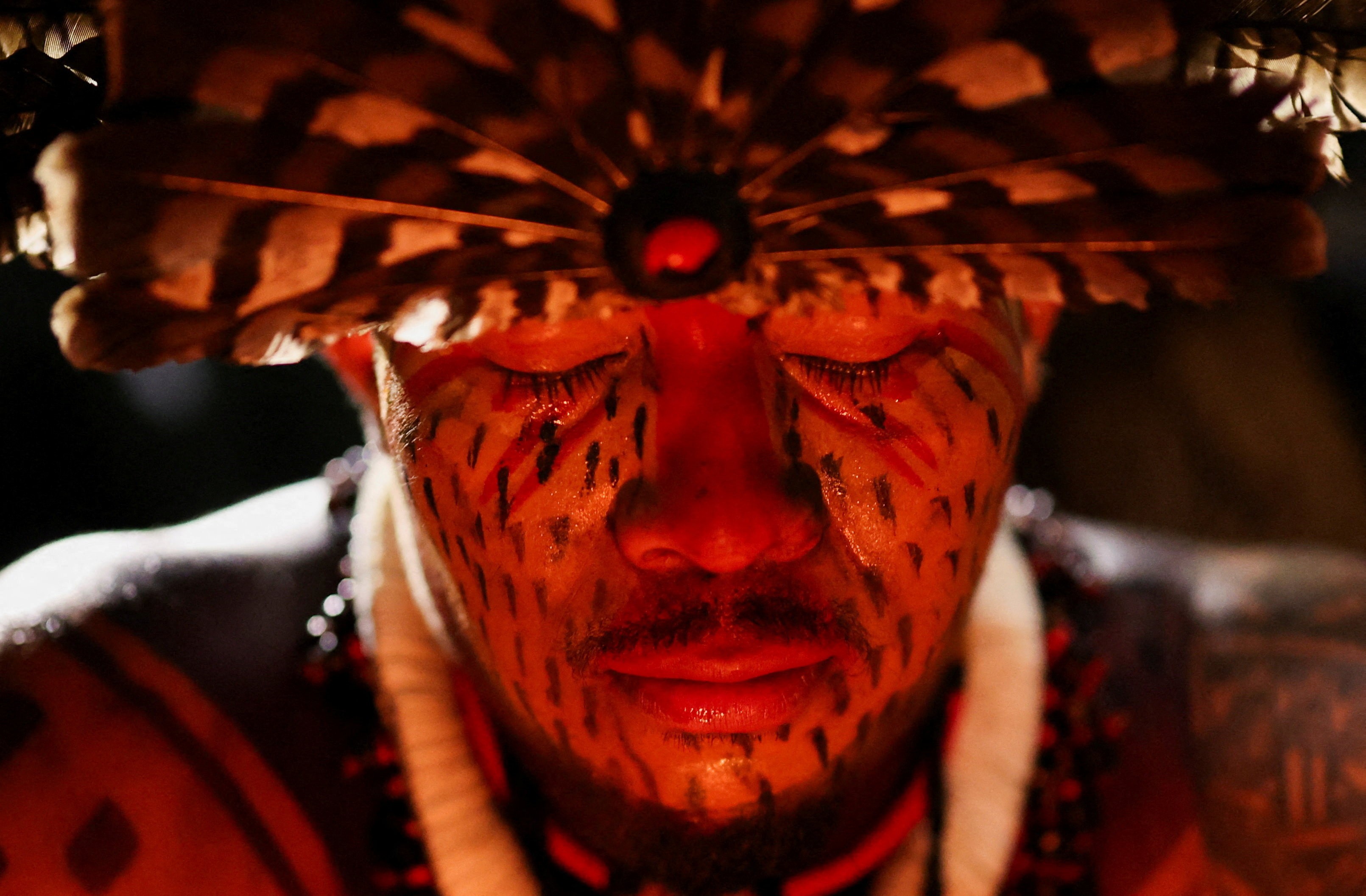 A close view of an Indigenous man wearing a traditional feathered headdress and makeup
