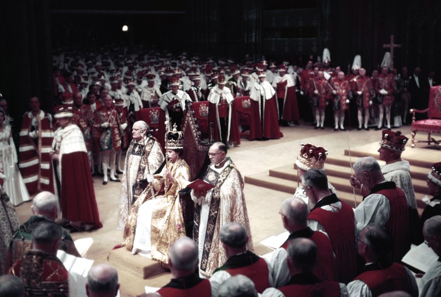 Hundreds attend a coronation ceremony, with the new queen seated on a throne.