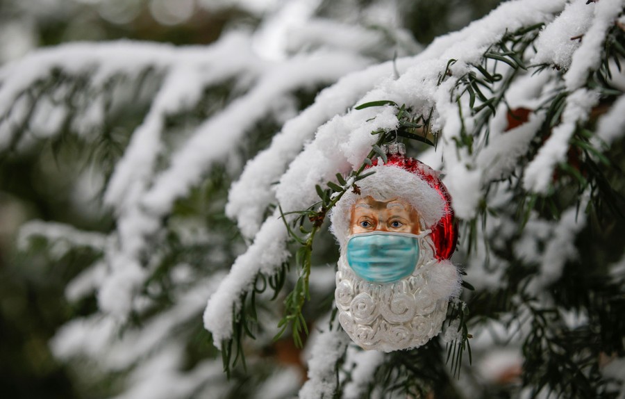 A Christmas ornament shaped as Santa Claus wearing a protective mask hangs from a snow-covered pine tree.