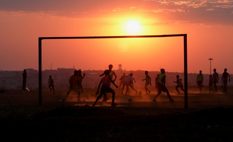 People play soccer on a dusty field at sunset.