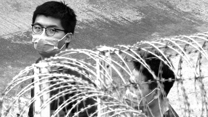 Two men wearing surgical masks walk behind a barbed-wire fence.