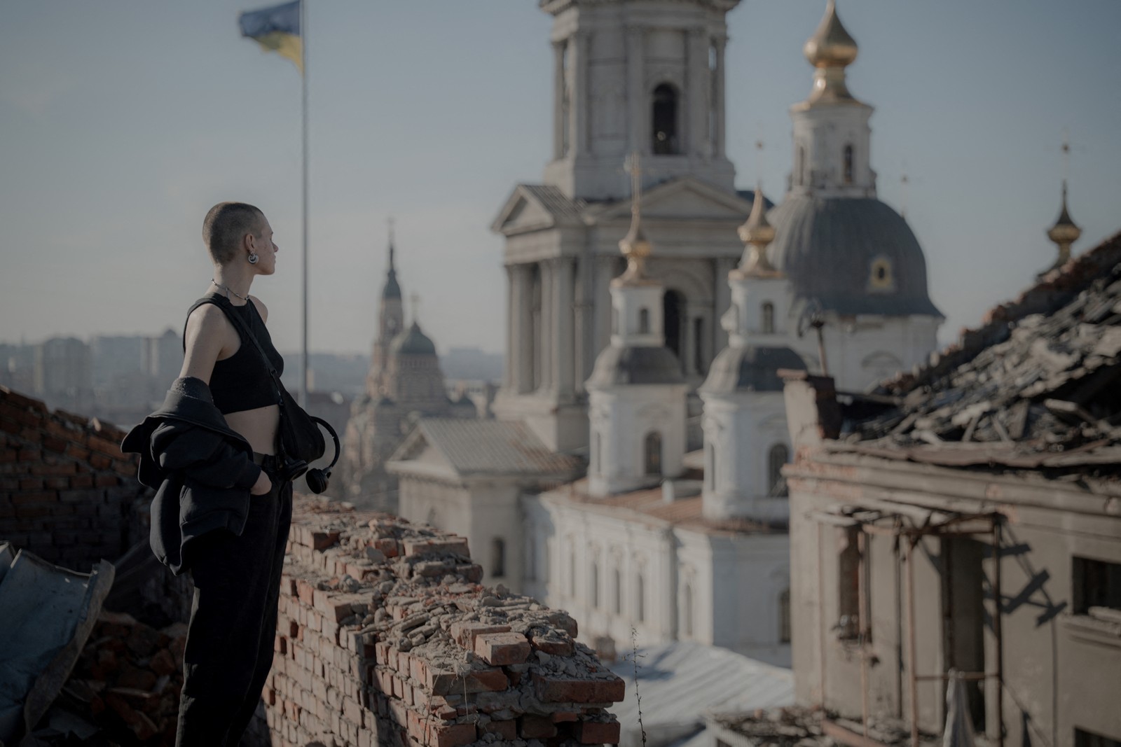 A civilian stands on a damaged rooftop overlooking a church and other large damaged buildings.