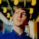 A photo of Alex Edelman surrounded by blurred lights outside of Hudson Theatre in New York