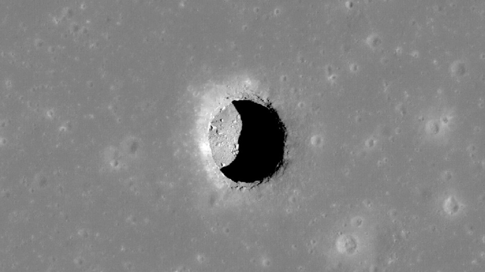 The opening to a lunar pit forms a black crescent on a gray landscape.