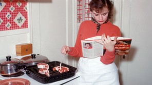A woman reads a cookbook next to meat cooking on the stove.