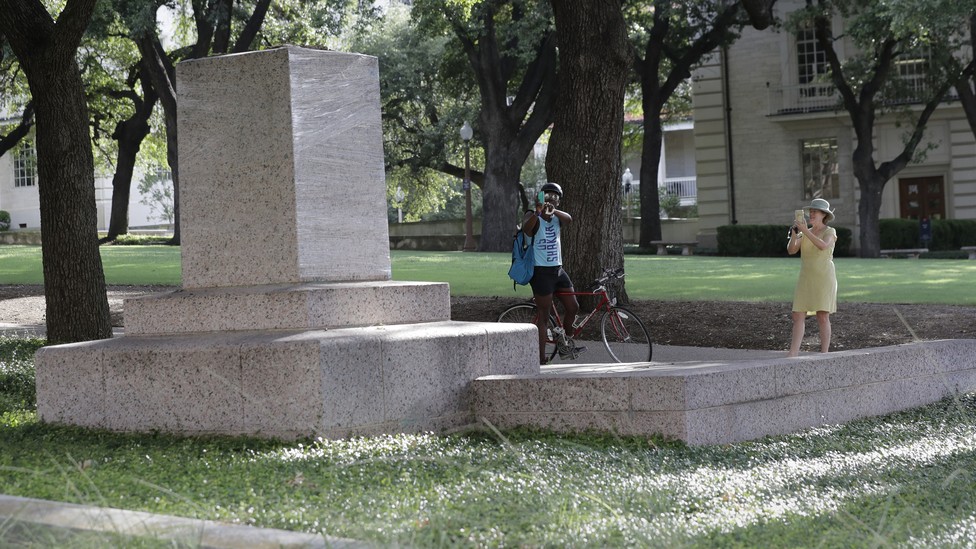 Two passersby take photos of a pedestal where a statue once stood.