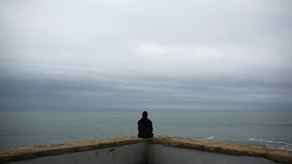 A person sitting on a low wall looking at the ocean under an overcast sky