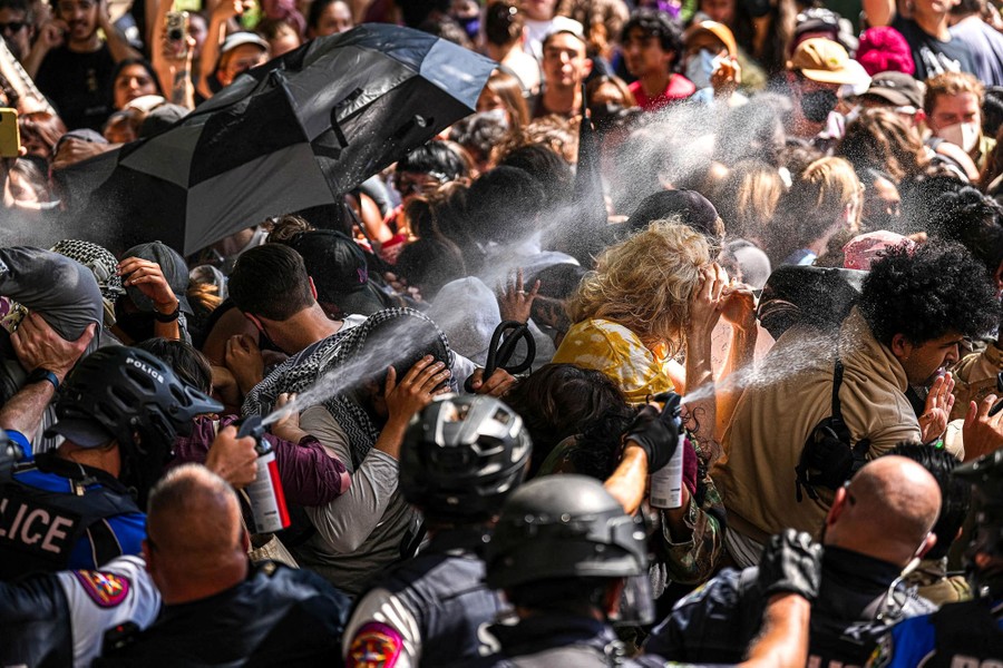 Several riot police officers are seen crowding against a large group of protesters, as at least two officers spray pepper spray into the crowd.