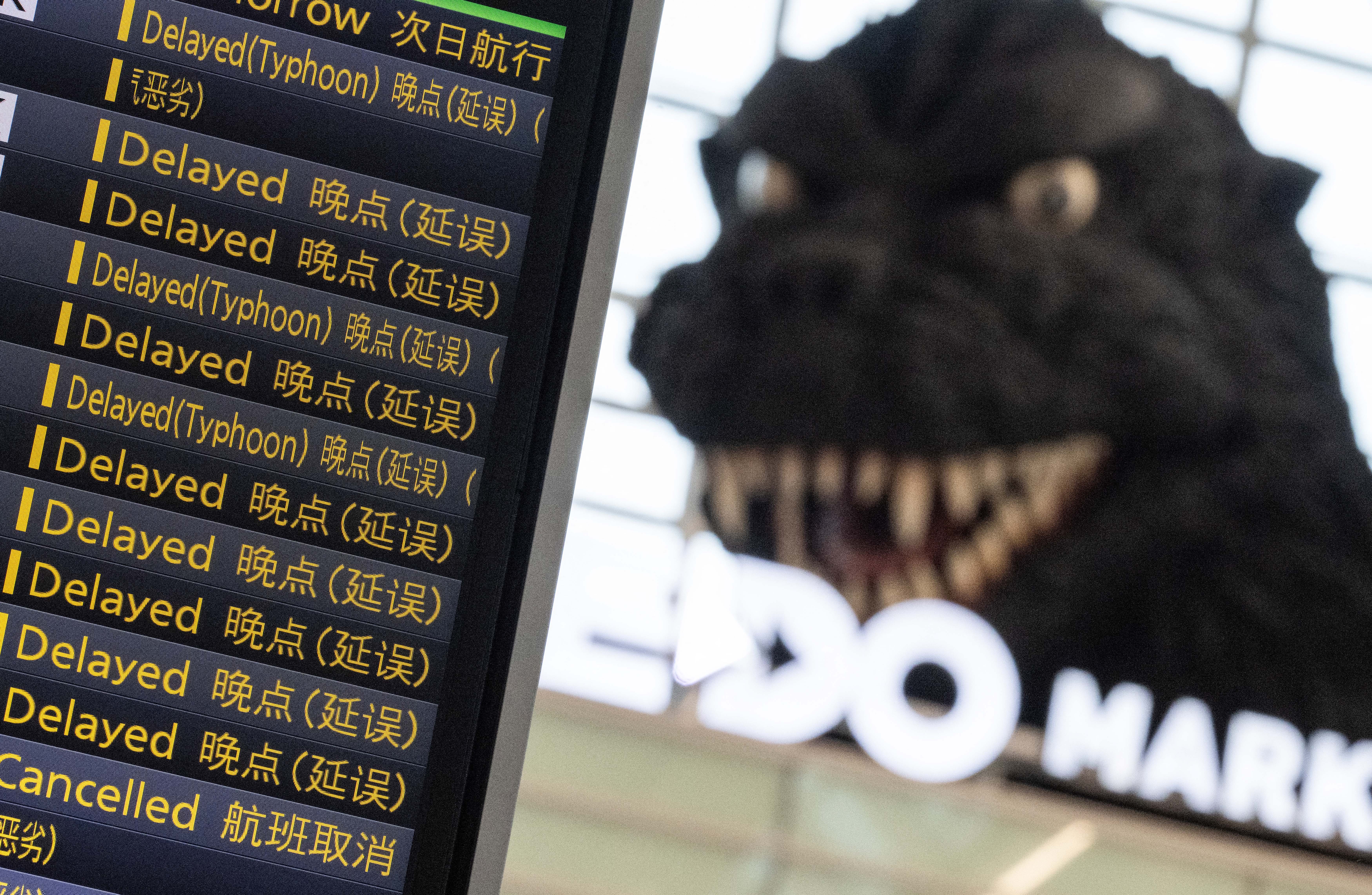 A flight board in an airport shows many cancelled or delayed flights, with a large Godzilla sculpture looming in the background.