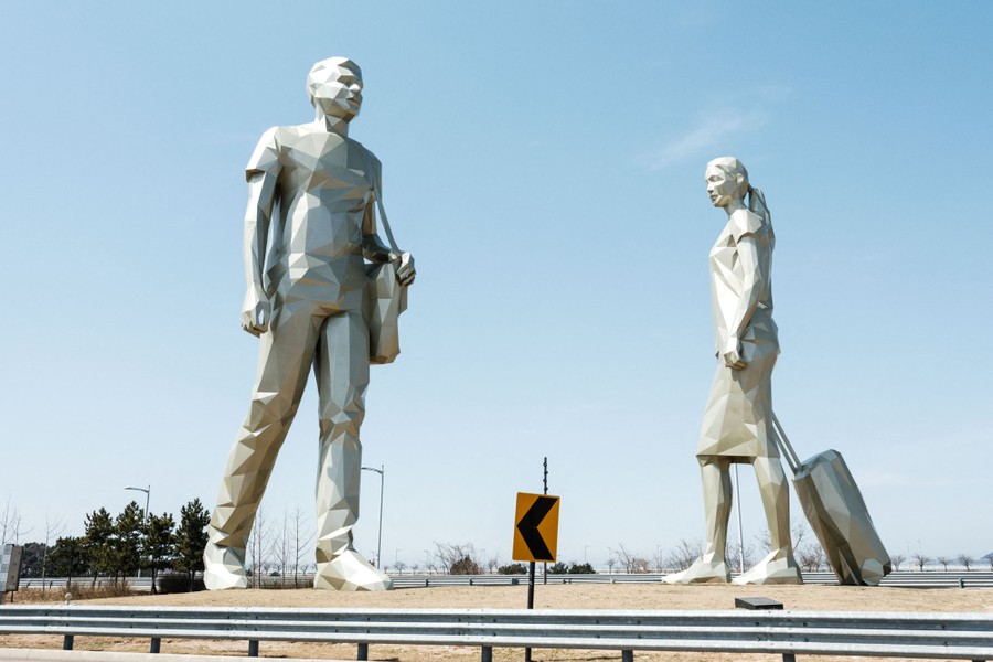 Two tall silvery sculptures depict people carrying luggage, set alongside a road near an airport.