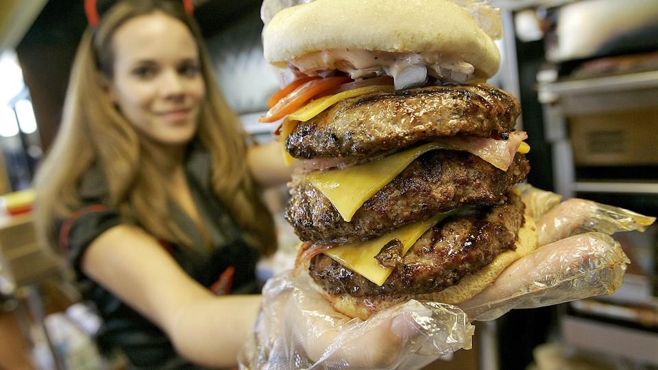 A restaurant worker in a red and black uniform holds a three-patty cheeseburger.