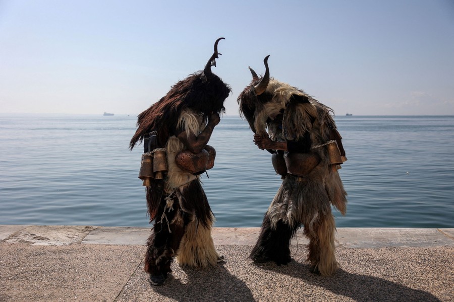 Two people stand on concrete near the sea, wearing costumes made of animal skins.