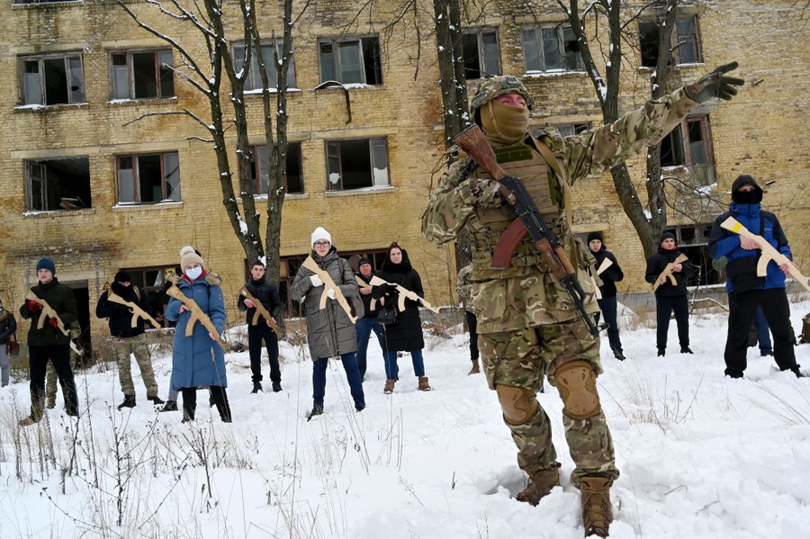 A military instructor holding a rifle stands before more than a dozen civilians in a snowy field in front of an empty building. Each student holds a mock rifle made of wood.