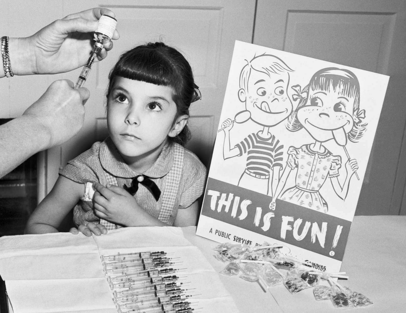 A young girl looks up as someone off camera prepares a syringe with a vaccine shot. The girl sits next to a sign with a drawing of a boy and girl holding hands and enjoying lollipops, and the the phrase 
