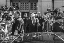 Black-and-white photo of Bob Menendez walking to a car from a building, his head down, flanked by photographers and law enforcement