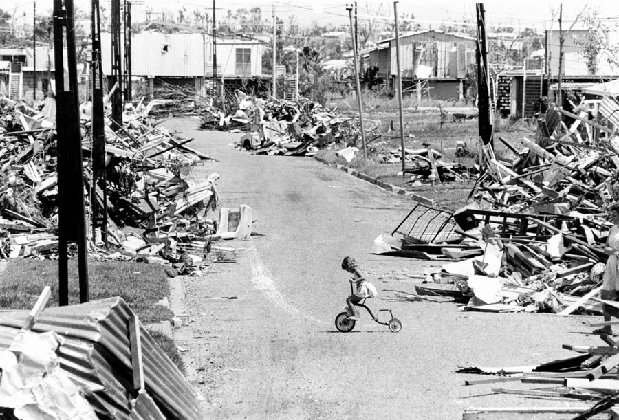 A young girl rides her tricycle on a street in the aftermath of a cyclone, with the debris of destroyed houses everywhere.